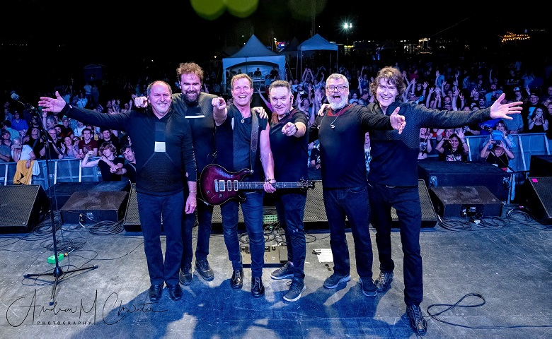  Six Strange Advance band members posing on outdoor stage at night, with audience in background