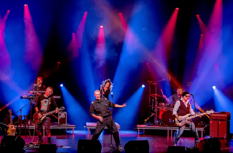 Six musicians from The Box band on stage, singing and playing instruments with dramatic stage lighting